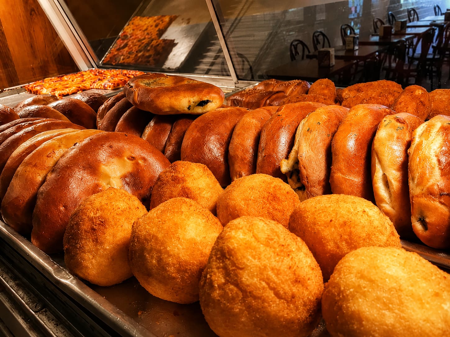 A counter display case packed with rows of golden calzones and arancini, with the dining room visible through the glass behind it.