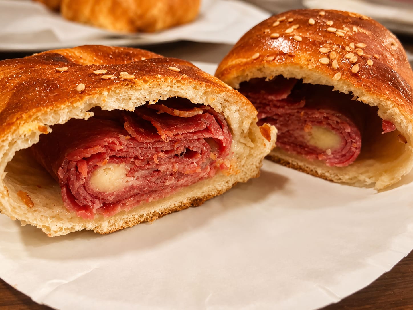 A sesame-topped roll cut open to show layers of cured meat and cheese, on a sheet of paper.