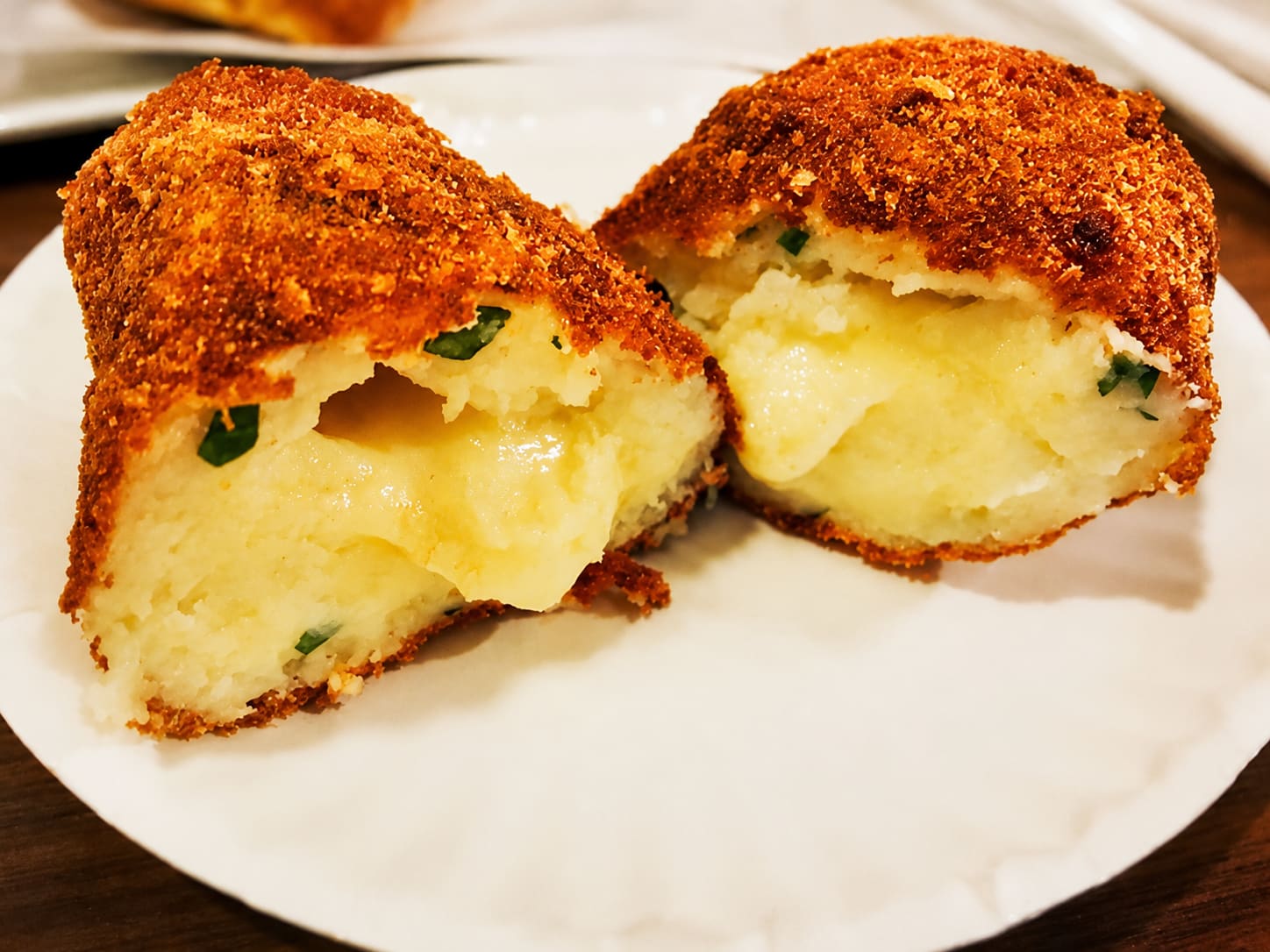 Two fried potato croquettes broken open on a plate, showing a soft cheesy potato filling flecked with herbs.