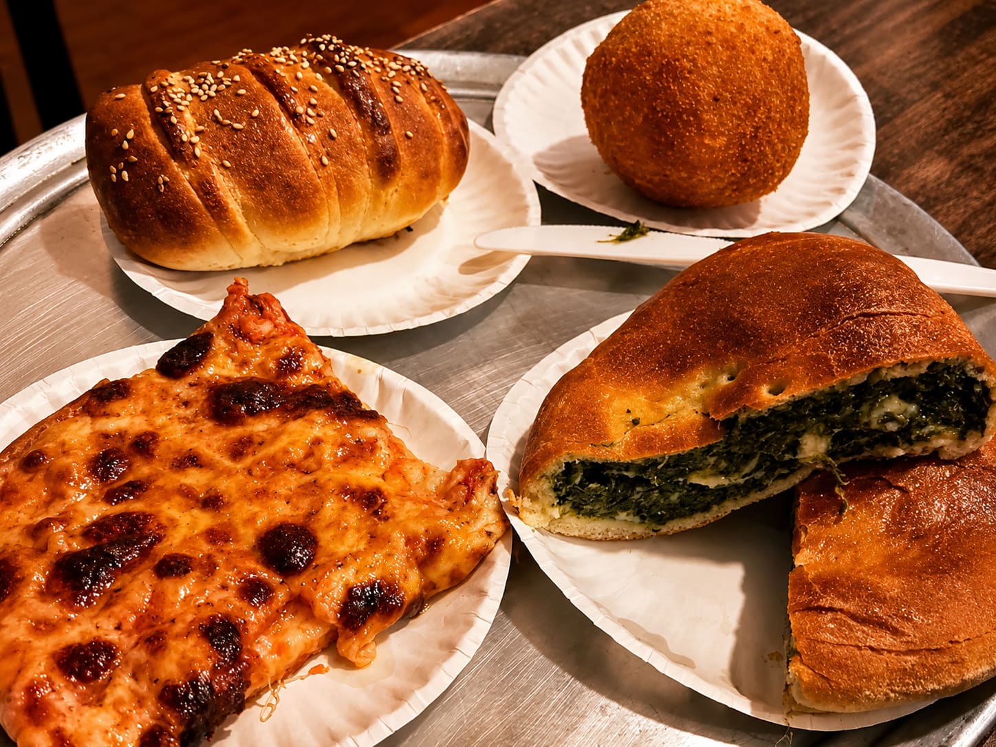 A steel counter with four paper plates on it — a sesame panino, an arancino, a square of Sicilian pizza and a spinach calzone cut open.